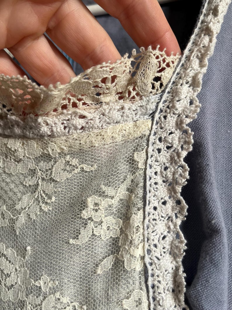 May include: Close-up of a delicate, off-white lace fabric. The lace features intricate floral patterns and is trimmed with a scalloped edge. The fabric appears to be part of a garment, possibly a vintage piece.
