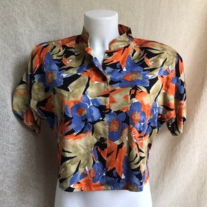 May include: A short-sleeved blouse with a vibrant floral pattern. The shirt features a mandarin collar and a button-down front. The floral design includes orange, blue, and black flowers with green and tan leaves. The blouse is displayed on a white mannequin.