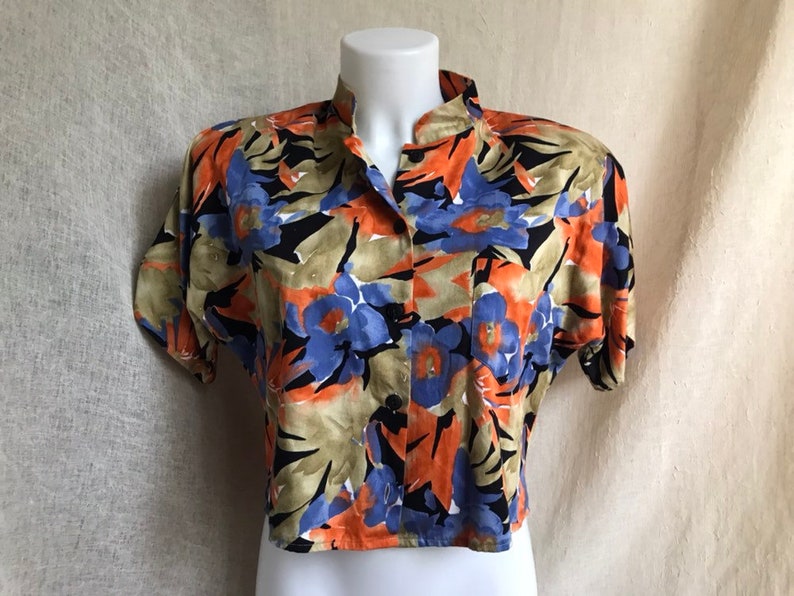May include: A short-sleeved blouse with a vibrant floral pattern. The shirt features a mandarin collar and a button-down front. The floral design includes orange, blue, and black flowers with green and tan leaves. The blouse is displayed on a white mannequin.