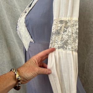 May include: A vintage-style dress in a soft periwinkle blue with white lace trim and sleeves. The sleeves are long and feature lace inserts. The dress has a V-neckline and a fitted bodice. A hand with a bracelet is touching the sleeve.
