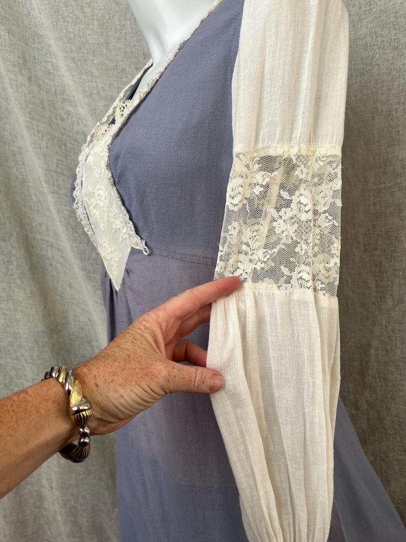 May include: A vintage-style dress in a soft periwinkle blue with white lace trim and sleeves. The sleeves are long and feature lace inserts. The dress has a V-neckline and a fitted bodice. A hand with a bracelet is touching the sleeve.
