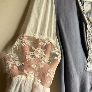 May include: A light blue dress with white sleeves and delicate lace cuffs. The dress features a V-neckline with white lace trim. The sleeves are a combination of white fabric and sheer lace, creating a romantic and vintage aesthetic.