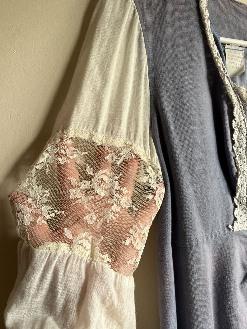 May include: A light blue dress with white sleeves and delicate lace cuffs. The dress features a V-neckline with white lace trim. The sleeves are a combination of white fabric and sheer lace, creating a romantic and vintage aesthetic.