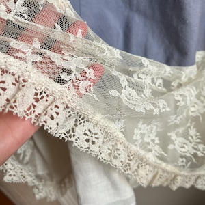 May include: Close-up of a vintage garment featuring delicate ivory lace trim. The lace is layered over a light blue fabric, creating a soft, textured effect. The intricate floral patterns of the lace are visible, adding a touch of elegance.