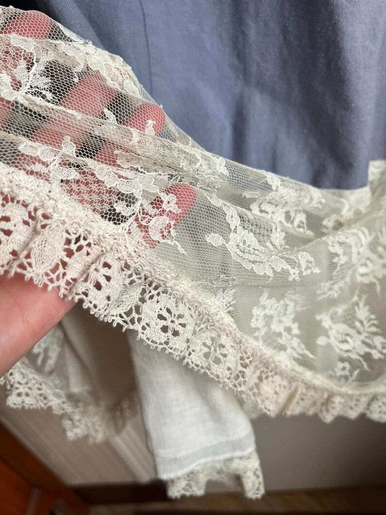May include: Close-up of a vintage garment featuring delicate ivory lace trim. The lace is layered over a light blue fabric, creating a soft, textured effect. The intricate floral patterns of the lace are visible, adding a touch of elegance.