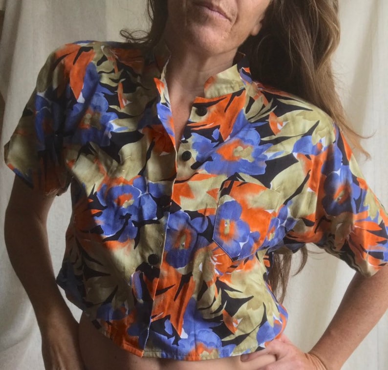 May include: A short-sleeved button-up shirt with a vibrant floral pattern. The shirt features a mandarin collar and a single chest pocket. The design includes orange, blue, and black floral elements on a tan background. The shirt is unbuttoned at the top.