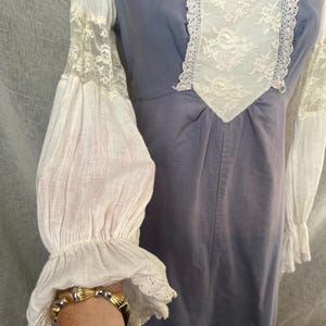 May include: A vintage-style dress in a muted blue hue, featuring white lace and fabric accents. The dress has long, billowing sleeves with lace detailing and a square neckline with lace trim. The overall design evokes a romantic, historical aesthetic.