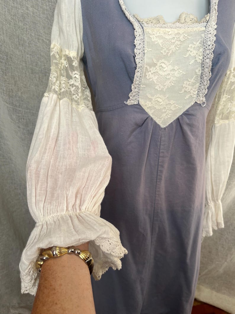 May include: A vintage-style dress in a muted blue hue, featuring white lace and fabric accents. The dress has long, billowing sleeves with lace detailing and a square neckline with lace trim. The overall design evokes a romantic, historical aesthetic.
