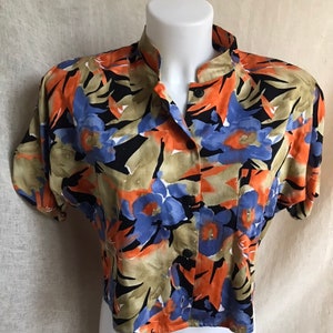 May include: A short-sleeved button-down shirt with a vibrant floral pattern. The shirt features a mandarin collar and a colorful design with orange, blue, black, and tan floral elements. The shirt is displayed on a white mannequin.