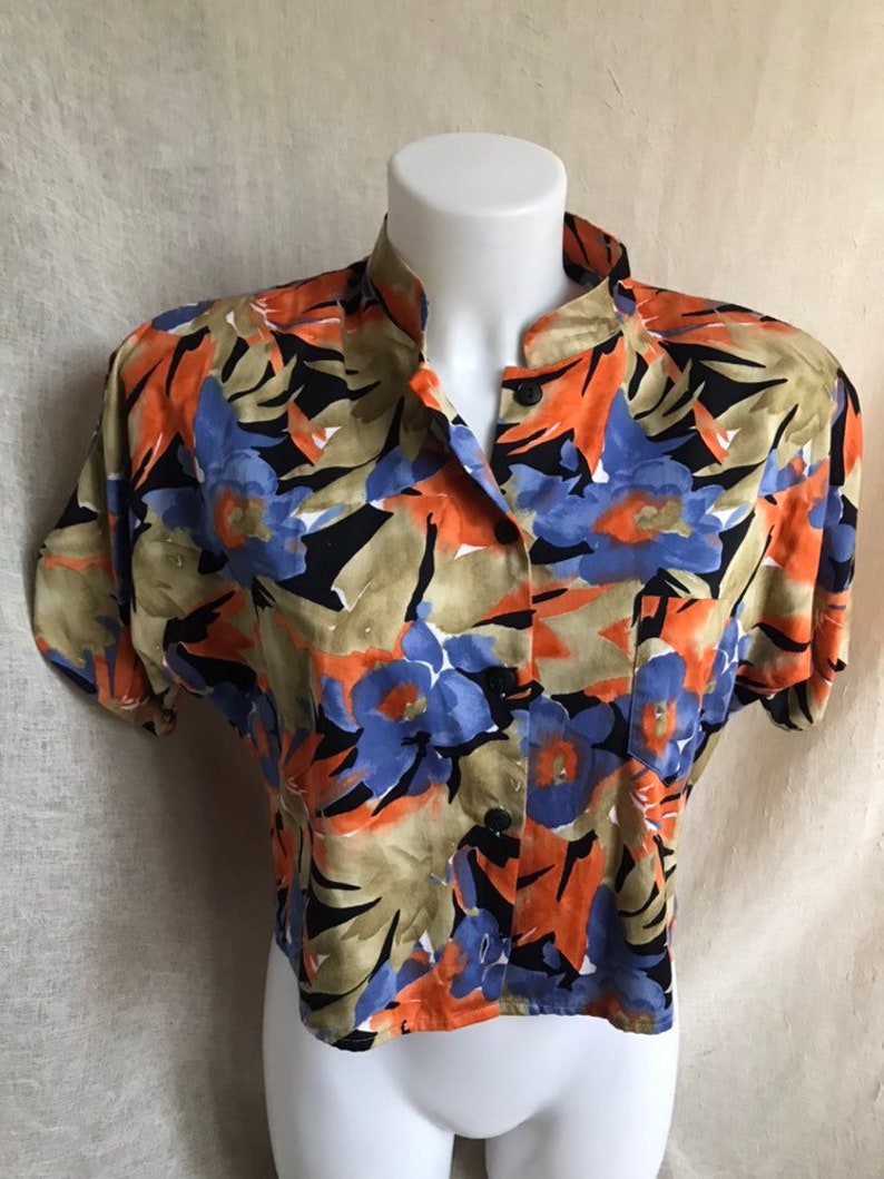May include: A short-sleeved button-down shirt with a vibrant floral pattern. The shirt features a mandarin collar and a colorful design with orange, blue, black, and tan floral elements. The shirt is displayed on a white mannequin.