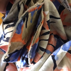 May include: Close-up of a beige fabric with a colorful floral pattern. The design includes orange, blue, and black shapes. The fabric is folded, revealing the edges and a tag that reads "brigitte".