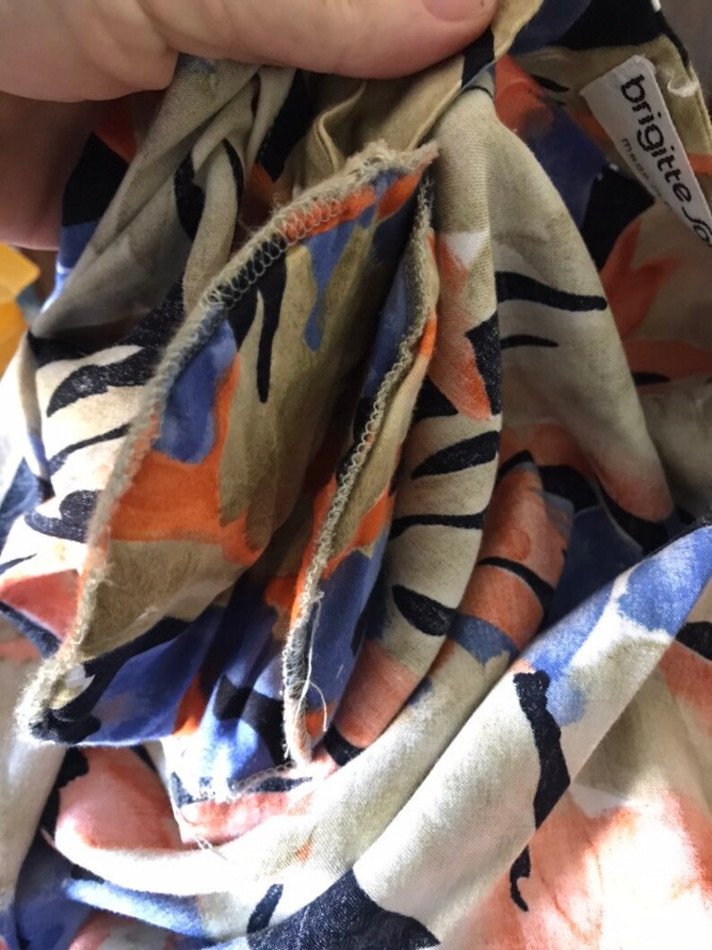 May include: Close-up of a beige fabric with a colorful floral pattern. The design includes orange, blue, and black shapes. The fabric is folded, revealing the edges and a tag that reads "brigitte".
