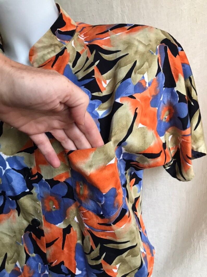 May include: A button-down shirt with a vibrant floral pattern in orange, blue, black, and tan. The shirt features a chest pocket and short sleeves. The fabric appears to be lightweight.