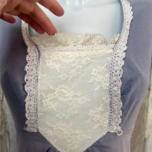 May include: A light blue dress with a square neckline and long sleeves. The bodice features a white lace panel and is trimmed with white lace. The dress is displayed on a mannequin.