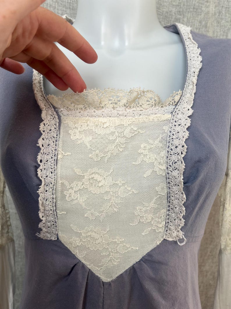 May include: A light blue dress with a square neckline and long sleeves. The bodice features a white lace panel and is trimmed with white lace. The dress is displayed on a mannequin.