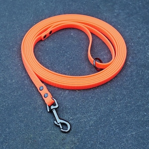 May include: A bright neon orange dog lead coiled in a circular shape. The lead features a smooth, flexible material with a black metal swivel snap hook at one end and a black metal D-ring attached near the handle loop, secured with black rivets.