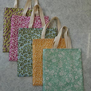 May include: Five fabric tote bags with cream-coloured handles. The bags display floral patterns in various colours, including green, pink, blue, yellow, and mint green. The bags are arranged diagonally, showcasing the different designs.
