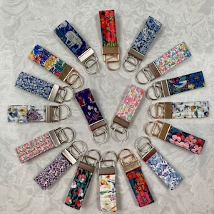May include: A collection of 18 keychains featuring various floral designs. Each keychain has a silver key ring and a gold-coloured metal loop. The keychains are arranged in a circle on a white background.