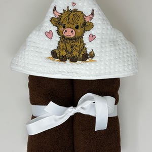 May include: A white hooded baby towel with a brown Highland cow embroidered on the hood, surrounded by pink hearts. The towel is paired with two rolled brown towels, tied with a white ribbon.