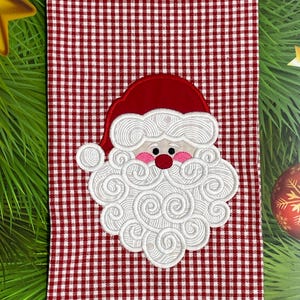 May include: A red and white checkered Christmas kitchen towel featuring an embroidered Santa Claus face. The Santa face has a red hat, pink cheeks, a red nose, and a white beard with swirling details. Festive holiday decor.