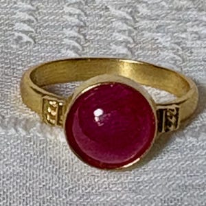 Può includere: Un anello d'oro con una grande pietra preziosa di rubino rotonda e cabochon. L'anello ha una fascia semplice con un design decorativo.