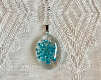 May include: A silver chain necklace with a clear oval pendant containing a teal blue pressed flower.