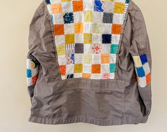 Puede incluir: Una chaqueta gris con un diseño de patchwork en la espalda. El patchwork está hecho de cuadrados de diferentes colores y patrones, incluyendo azul, verde, amarillo, naranja y rojo. La chaqueta tiene cuello y mangas largas.