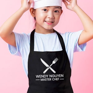 May include: A young person wearing a black apron with a white chef's hat. The apron has a white design of crossed utensils and the text "WENDY NGUYEN - MASTER CHEF - "