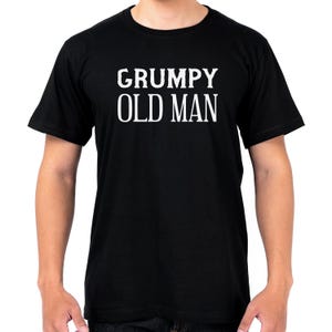 May include: Black crew neck t-shirt with the white text "GRUMPY OLD MAN" printed across the chest. The short-sleeved shirt is made of a soft material. The person wearing the shirt is standing.