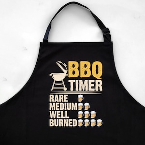 May include: Black BBQ apron with a yellow graphic that says "BBQ TIMER" and a grill with a thermometer. Below the grill, it says "RARE, MEDIUM, WELL, BURNED" with beer mugs representing each level of doneness.