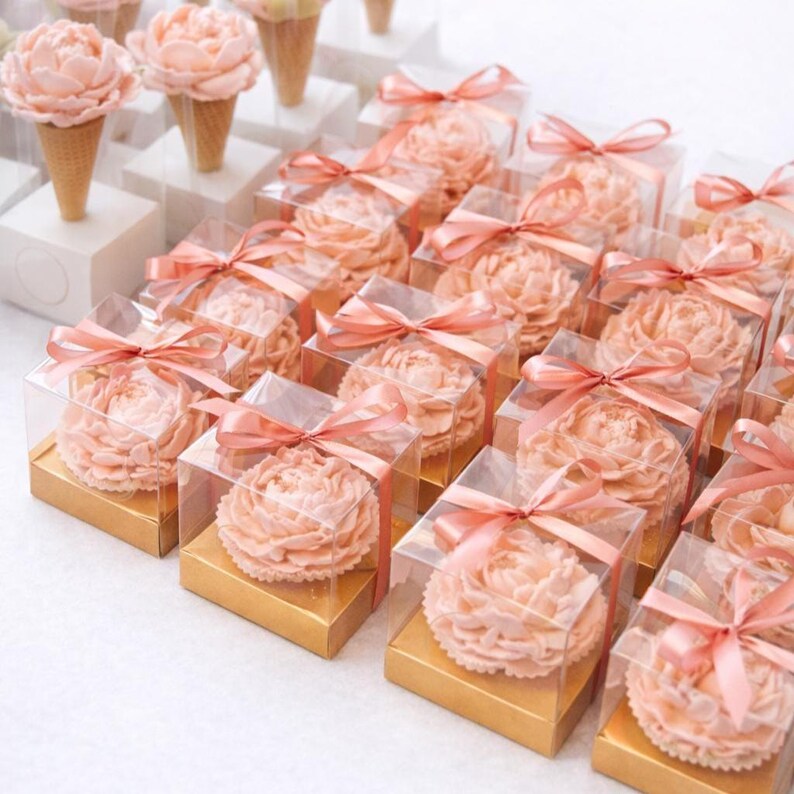 May include: Rows of peach-colored peony-shaped treats in clear boxes with peach ribbon bows. Some treats are on ice cream cones. The boxes sit on gold bases, arranged on a white surface. A delightful presentation for a special occasion.