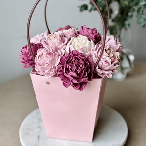 Op de afbeelding: Een roze papieren cadeauzak met een koperkleurige handgreep, gevuld met roze en paarse bloemen. De tas staat op een witte marmeren schaal. De bloemen zijn een mix van lichtroze, donkerroze en wit.