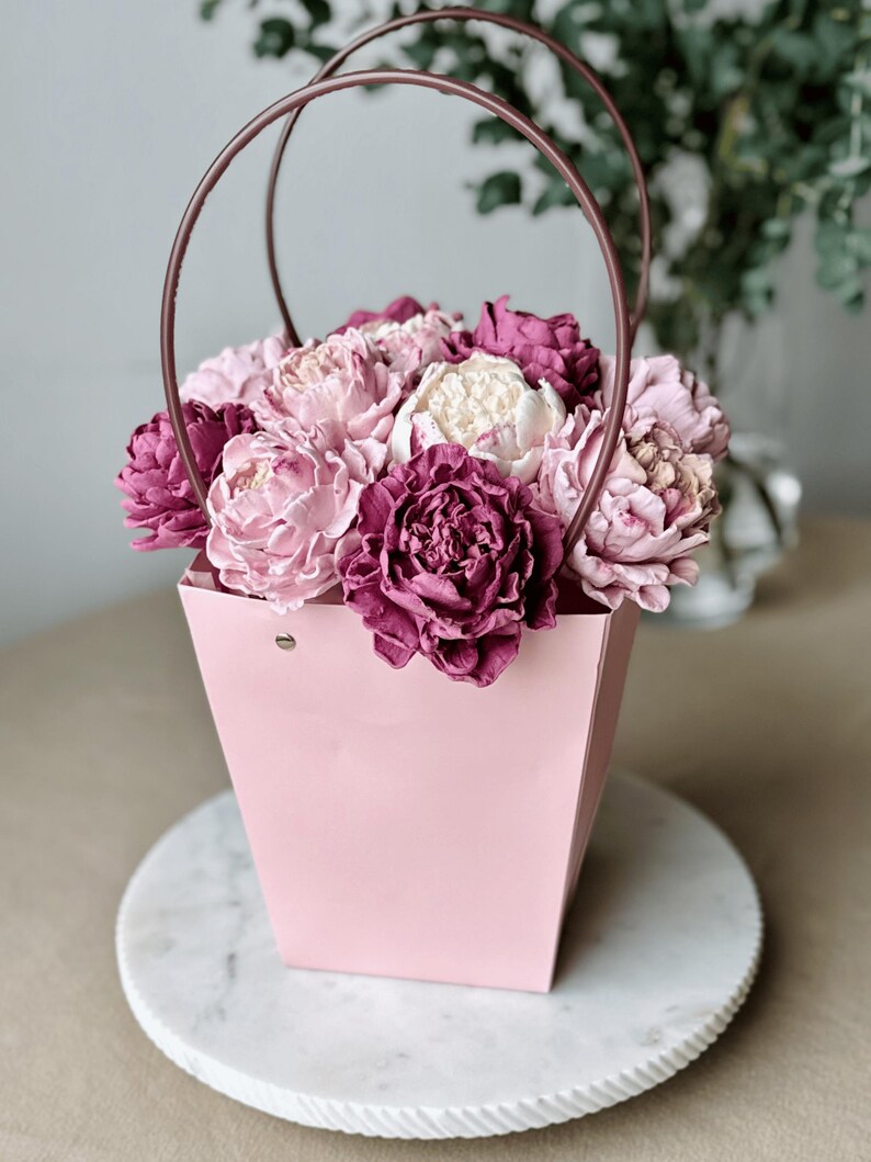Op de afbeelding: Een roze papieren cadeauzak met een koperkleurige handgreep, gevuld met roze en paarse bloemen. De tas staat op een witte marmeren schaal. De bloemen zijn een mix van lichtroze, donkerroze en wit.
