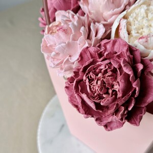 Op de afbeelding: Een roze, zeshoekige container gevuld met verschillende tinten roze en cr&egrave;mekleurige bloemen. De bloemen omvatten grote, getextureerde bloesems met gedetailleerde bloemblaadjes, waardoor een zachte, romantische esthetiek ontstaat. De container heeft een handvat.