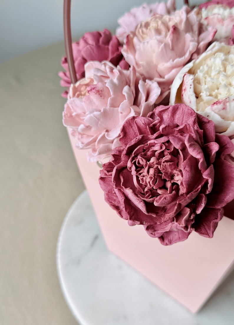 Op de afbeelding: Een roze, zeshoekige container gevuld met verschillende tinten roze en cr&egrave;mekleurige bloemen. De bloemen omvatten grote, getextureerde bloesems met gedetailleerde bloemblaadjes, waardoor een zachte, romantische esthetiek ontstaat. De container heeft een handvat.