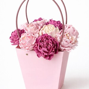 Op de afbeelding: Een roze papieren bloemenmand gevuld met een assortiment pioenrozen in tinten roze, cr&egrave;me en diep magenta. De mand heeft een bijpassende roze handgreep, perfect om cadeau te geven.