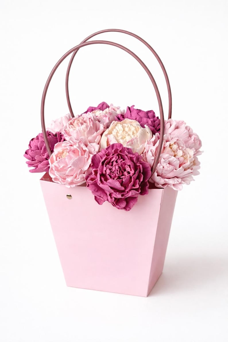 Op de afbeelding: Een roze papieren bloemenmand gevuld met een assortiment pioenrozen in tinten roze, cr&egrave;me en diep magenta. De mand heeft een bijpassende roze handgreep, perfect om cadeau te geven.