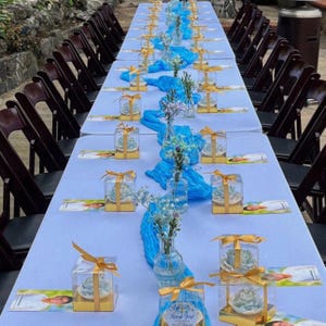 May include: A long table set for an event, covered with a light blue tablecloth. The table is decorated with blue fabric runners, small glass vases with flowers, and gift boxes tied with gold ribbons. Dark brown chairs are arranged around the table.