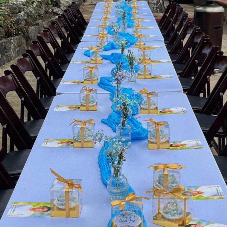 May include: A long table set for an event, covered with a light blue tablecloth. The table is decorated with blue fabric runners, small glass vases with flowers, and gift boxes tied with gold ribbons. Dark brown chairs are arranged around the table.