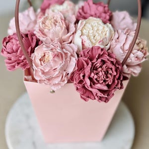 Op de afbeelding: Een roze papieren cadeauzakje gevuld met een assortiment roze en cr&egrave;mekleurige pioenrozen. De bloemen hebben verschillende roze tinten, van licht tot dieproze. De tas heeft een bijpassende handgreep.