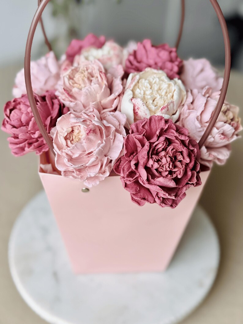 Op de afbeelding: Een roze papieren cadeauzakje gevuld met een assortiment roze en cr&egrave;mekleurige pioenrozen. De bloemen hebben verschillende roze tinten, van licht tot dieproze. De tas heeft een bijpassende handgreep.