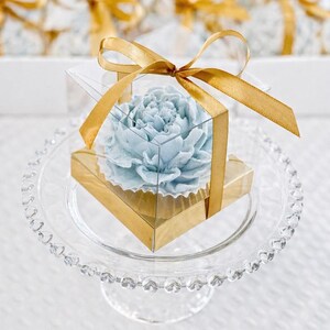 May include: A light blue, flower-shaped item is encased in a clear, square box with a gold base. The box is tied with a gold ribbon and sits on a clear glass cake stand with a beaded edge. The background is blurred, suggesting a display.