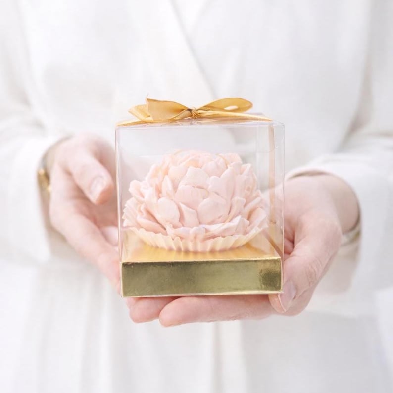 May include: A light pink peony flower in a clear square box with a gold base and a gold ribbon. The flower is the focal point, with detailed petals. The box is held by a person wearing a white garment.
