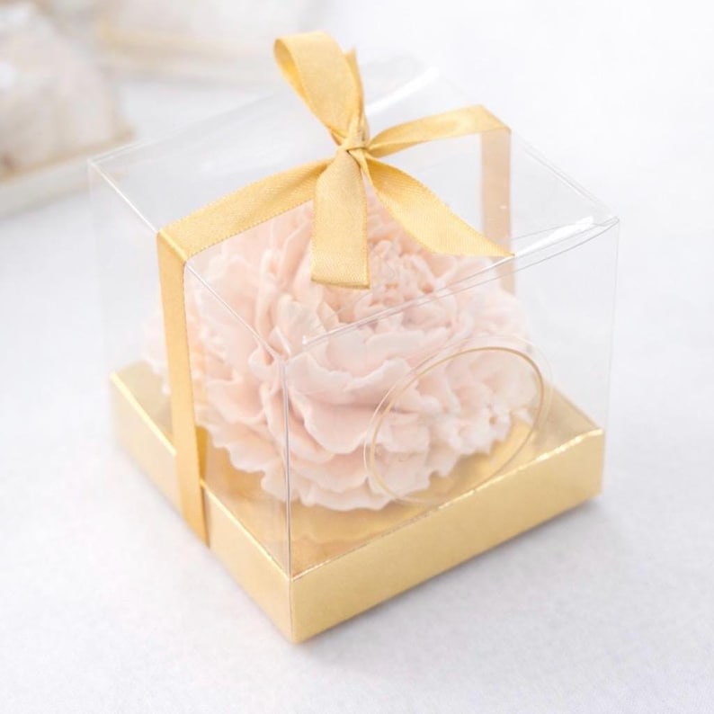 May include: A light pink flower is encased in a clear, square box with a gold base. A gold ribbon is tied in a bow on top. The flower is the focal point, with delicate petals. The box is a gift box.