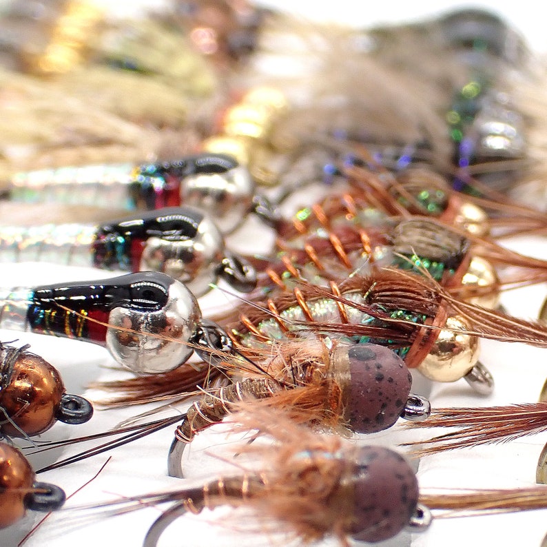 May include: A close-up of a variety of fly fishing lures with different colors, materials, and designs. The lures are arranged in a row on a white surface.