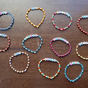 May include: Twelve colorful beaded bracelets with different names spelled out on them. The bracelets are made with small, colorful beads and are arranged in two rows on a wooden surface.