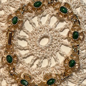 May include: A gold bracelet with green stones set in a decorative design. The bracelet is on a white crocheted surface.
