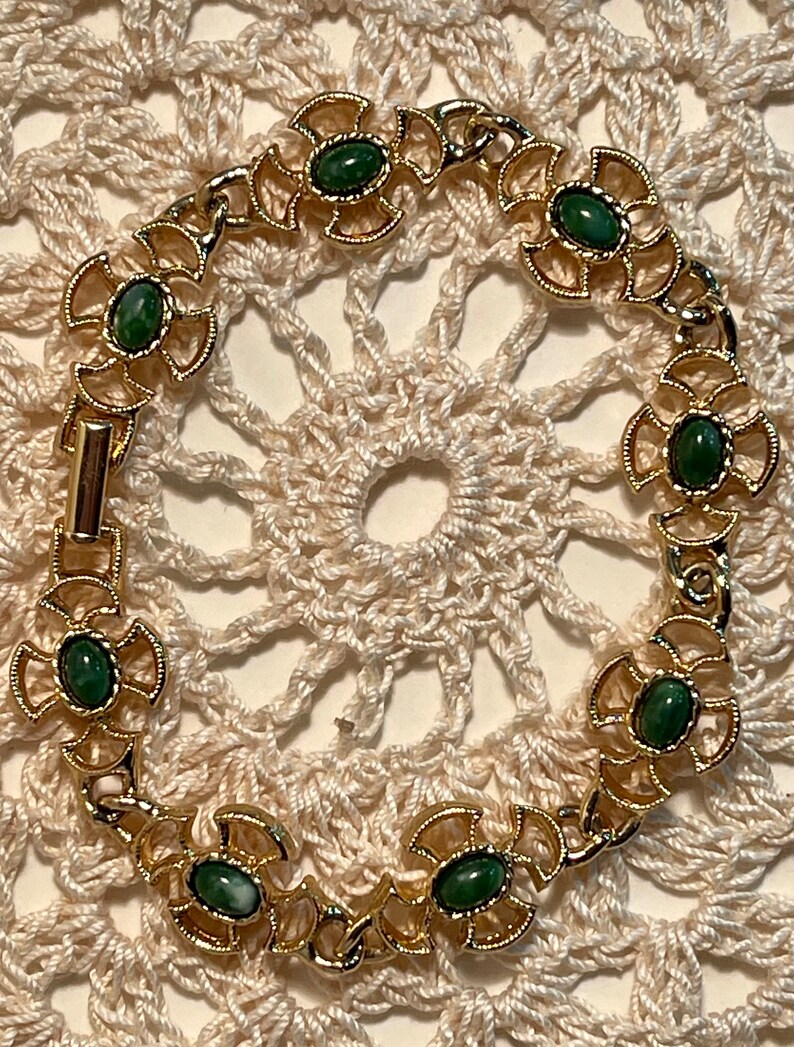 May include: A gold bracelet with green stones set in a decorative design. The bracelet is on a white crocheted surface.