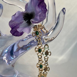 May include: A gold chain bracelet with green stone accents. The bracelet is on a clear plastic hand display.