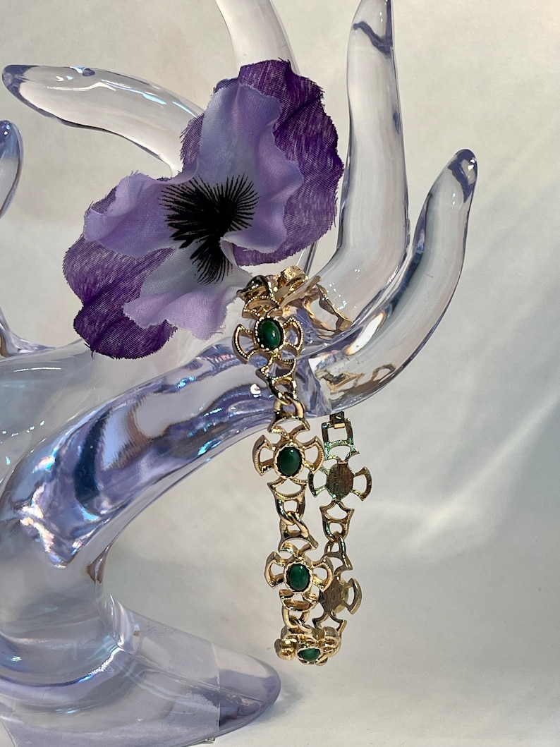 May include: A gold chain bracelet with green stone accents. The bracelet is on a clear plastic hand display.